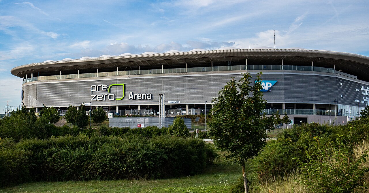 The PreZero Arena from outside: a low grey stadium with a perforated metal skin and a scalloped white roof edge, the PreZero Arena name on the facade and a large SAP logo beside it, hedges and young trees in front