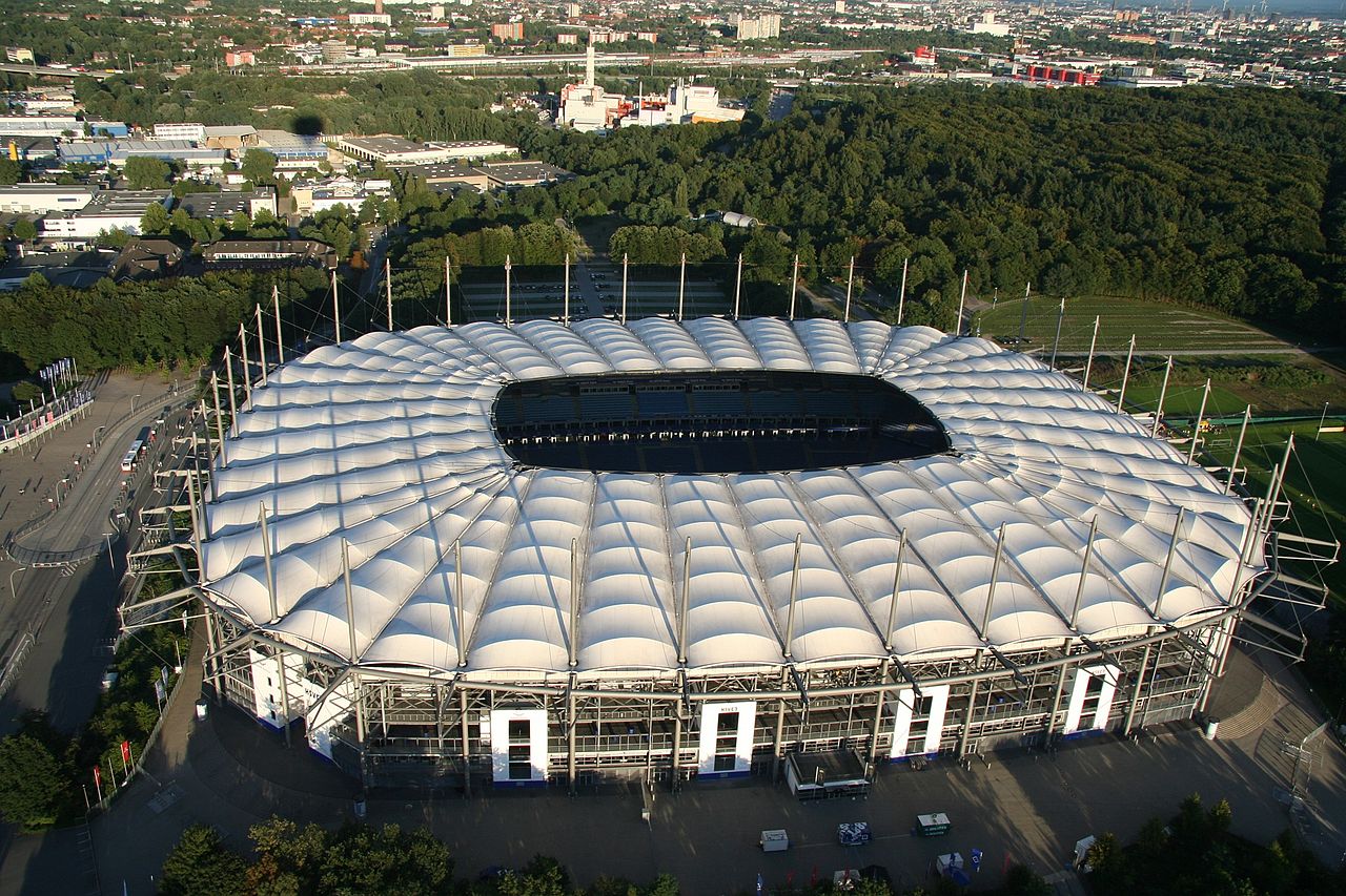 The Volksparkstadion from the air: an oval stadium with a white tented roof of inflated panels held on slender masts, surrounded by dense woodland with the city beyond