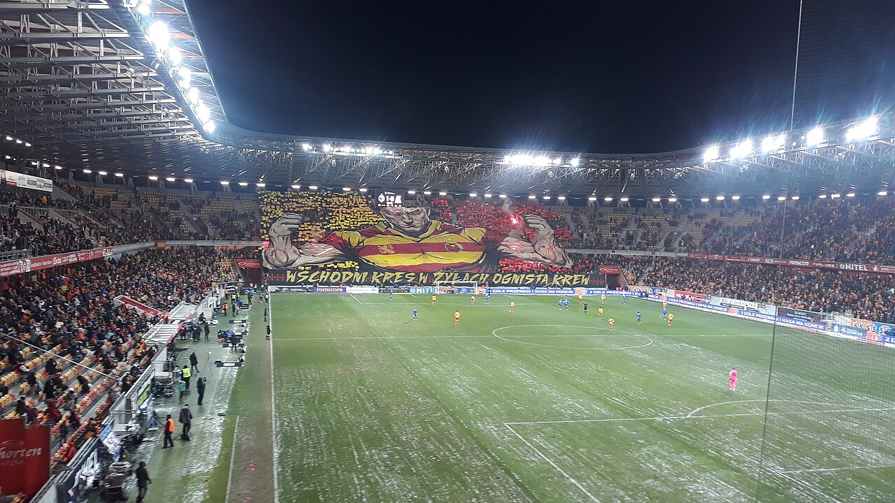 A packed Białystok stadium under floodlights at night, the home end covered by a huge painted banner of a bare-chested figure in Jagiellonia's yellow and red stripes