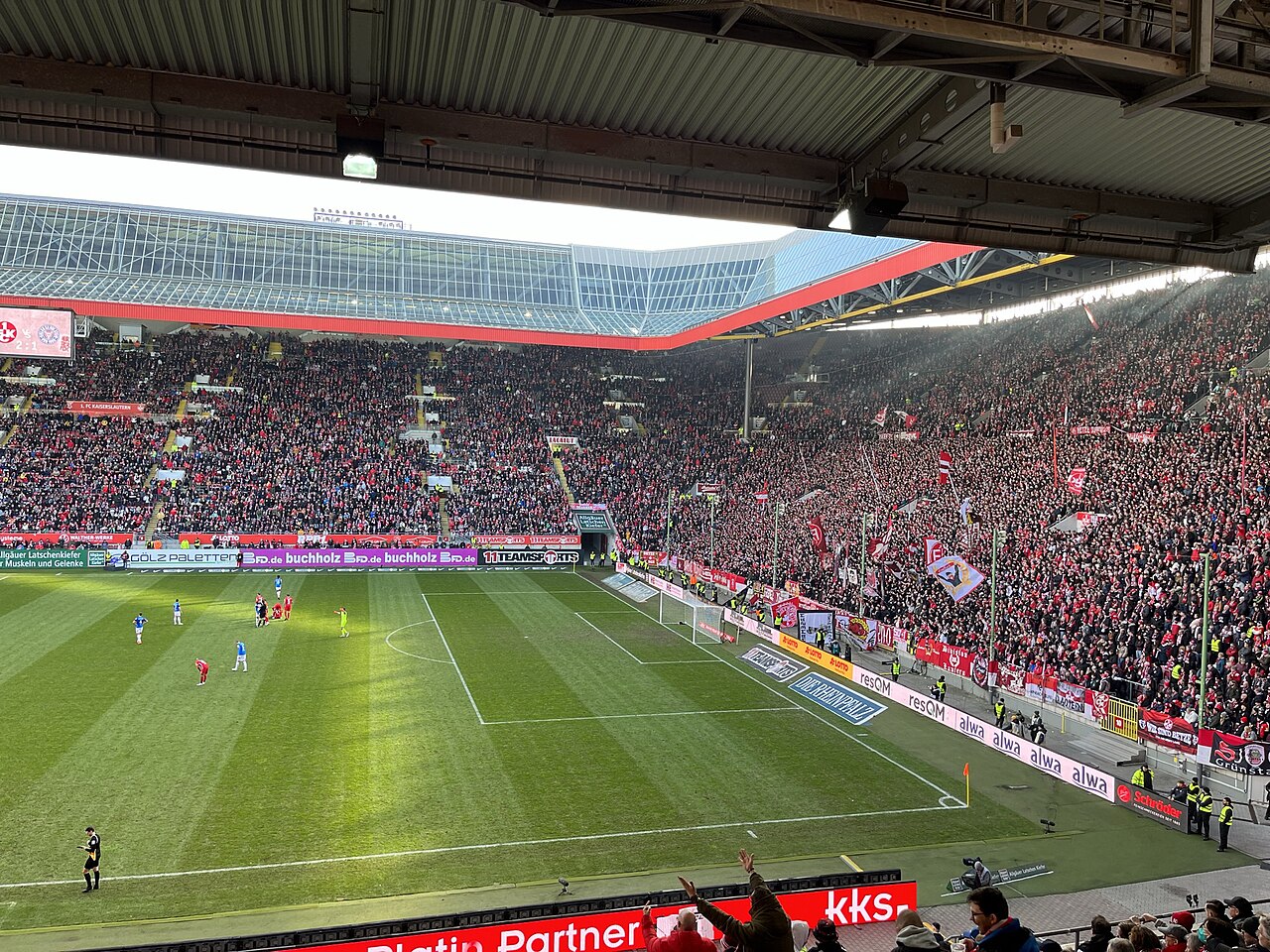 Inside a full Fritz-Walter-Stadion during a league match: steep stands packed with supporters in red, a large standing terrace behind the goal waving flags, and a roof edge of red steel and glass above them