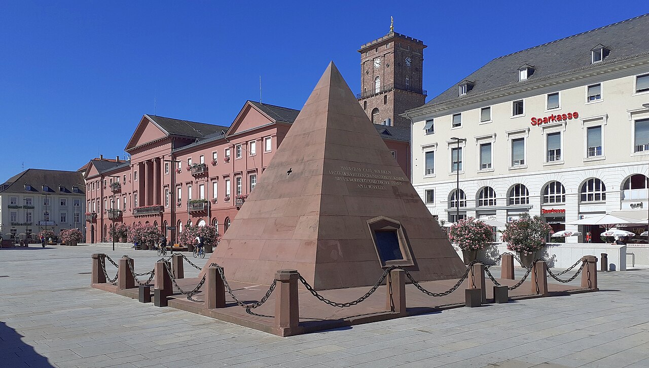 The market square in Karlsruhe: a red sandstone pyramid about the height of a house standing on the paved square behind a low chain fence, with the pink classical town hall and its clock tower on one side and a white arcaded building on the other