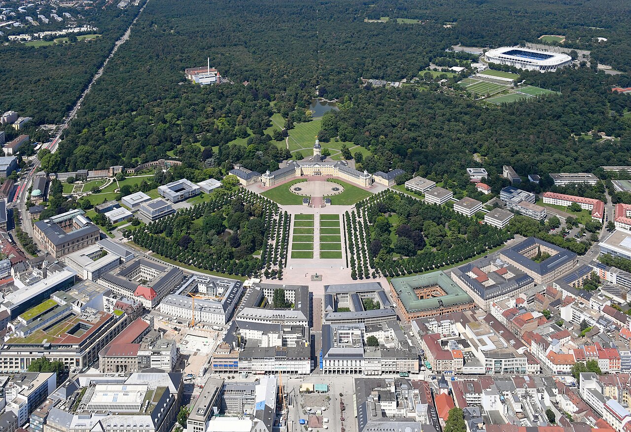 Karlsruhe from the air: the long yellow palace at the centre with avenues of trees and paved boulevards radiating outwards from it like the ribs of a fan, the dense city below and the wooded Hardtwald above, with the white roofs of the football stadium visible among the trees in the top right corner