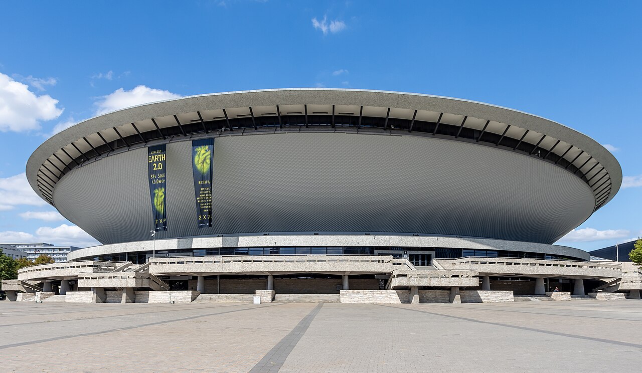 Spodek in Katowice: a large concrete arena shaped like a flying saucer, its saucer roof overhanging a ring of angled supports above a wide paved plaza