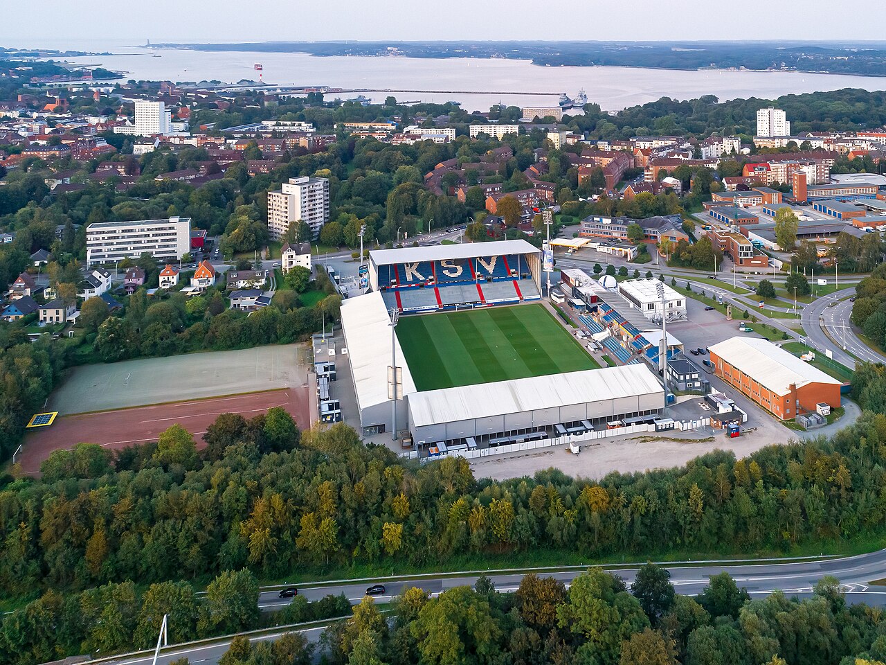 The Holstein-Stadion from the air: a small ground with one covered main stand whose blue and red seats spell out KSV, low stands on the other sides, woodland along one edge, the roofs of Kiel beyond and the Kiel fjord with a grey warship on it in the distance
