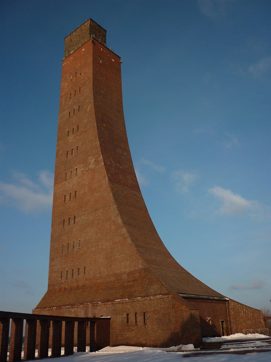 The naval memorial at Laboe: a tall red-brick tower with a sweeping curved buttress rising from a low brick hall, lit by low sun against a deep blue sky, with snow on the ground around it