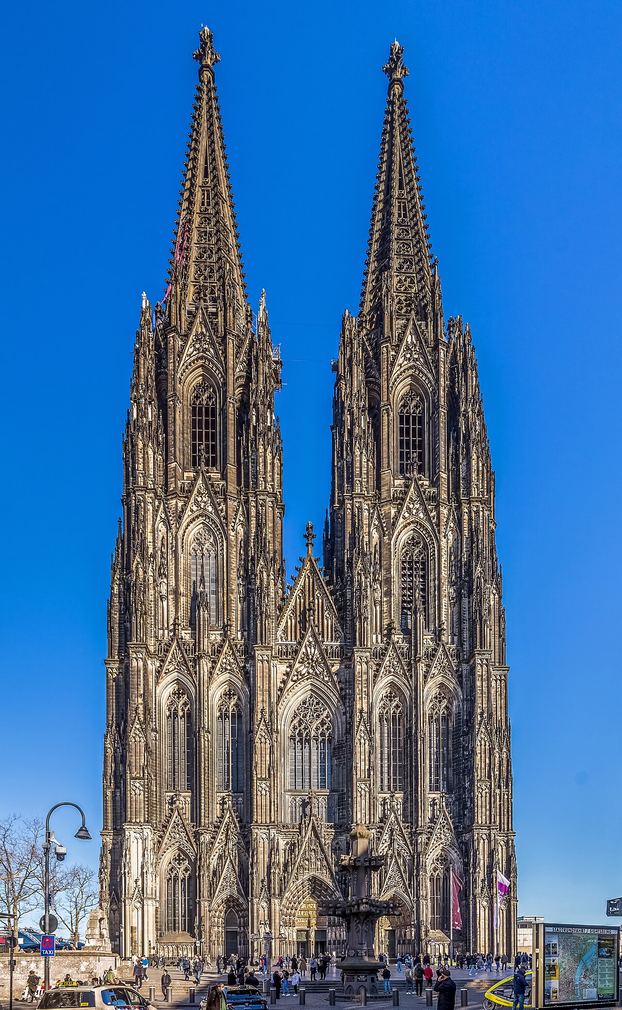 The west front of Cologne Cathedral: two enormous blackened Gothic spires rising side by side above a deeply carved facade, with people crossing the square in front of it under a clear blue sky