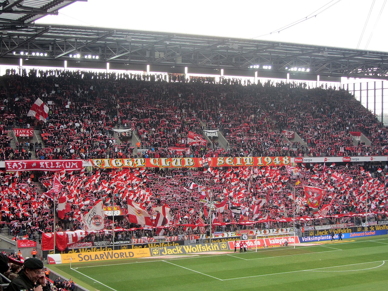 The packed home end at the RheinEnergieStadion: thousands of supporters waving red and white flags beneath a long banner reading ECHT CLUB SEIT 1948, with the covered upper tier above