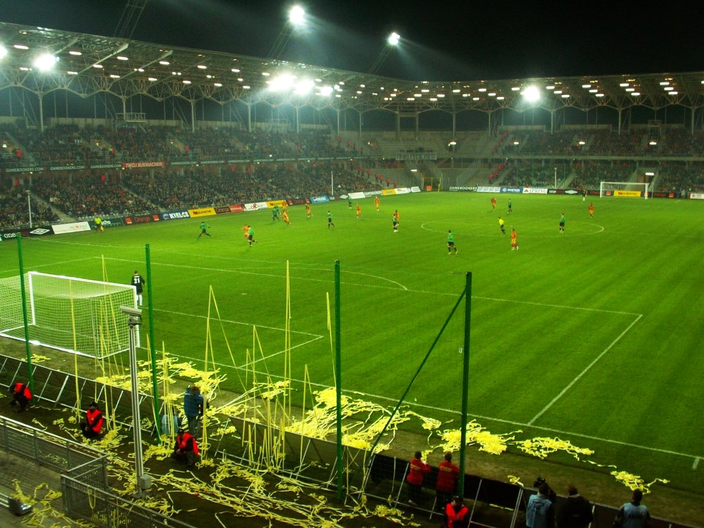 A floodlit night match at Korona Kielce seen from a corner of the stand: full tiers under a lit roof, players on a bright green pitch, and yellow paper streamers thrown across the running-track-free touchline