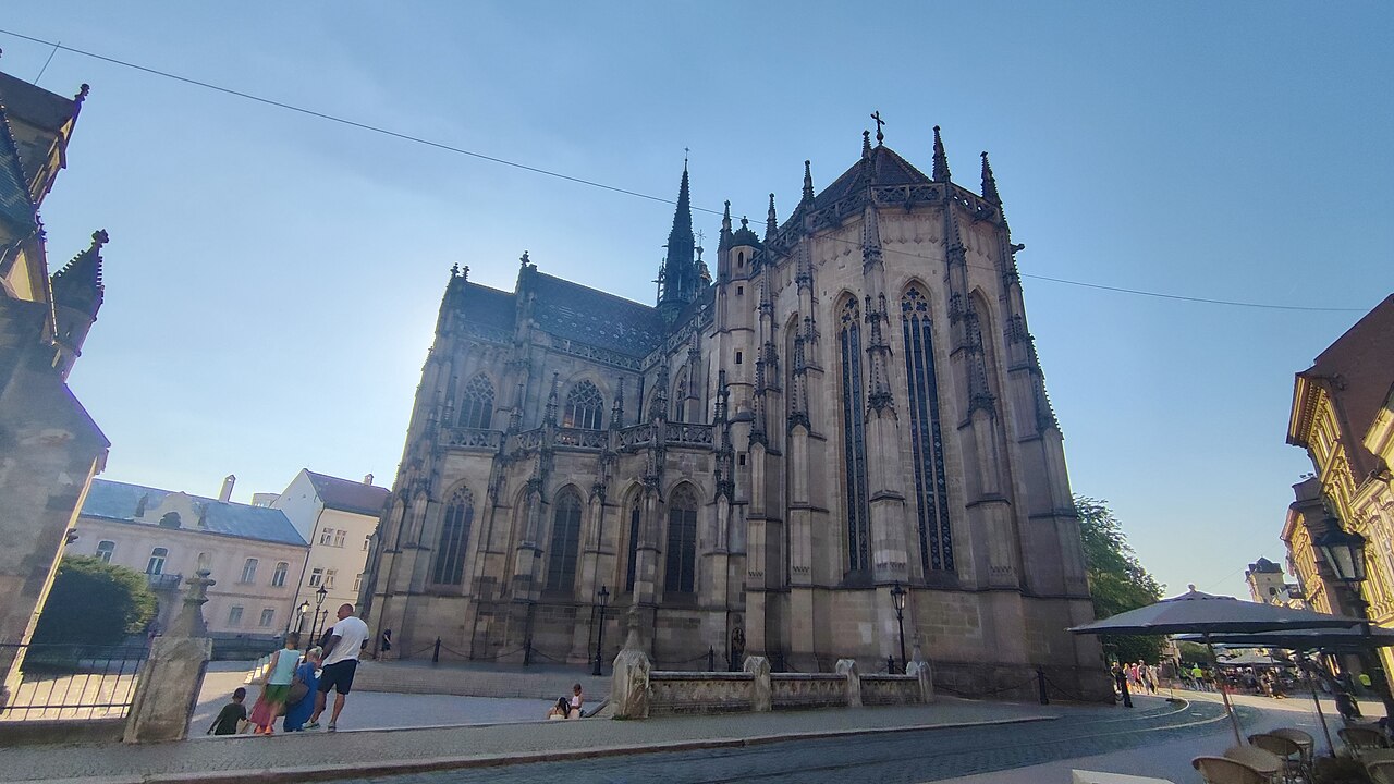 St Elisabeth's Cathedral in Košice seen from the side: a large Gothic church of pale stone with steep gables, pinnacles and a slender spire, standing on a paved street with cafés alongside