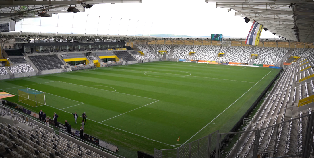 Inside the Košická futbalová aréna: a closed bowl of black, white and yellow seats under a continuous white roof, an empty green pitch, and low hills visible above the far stand