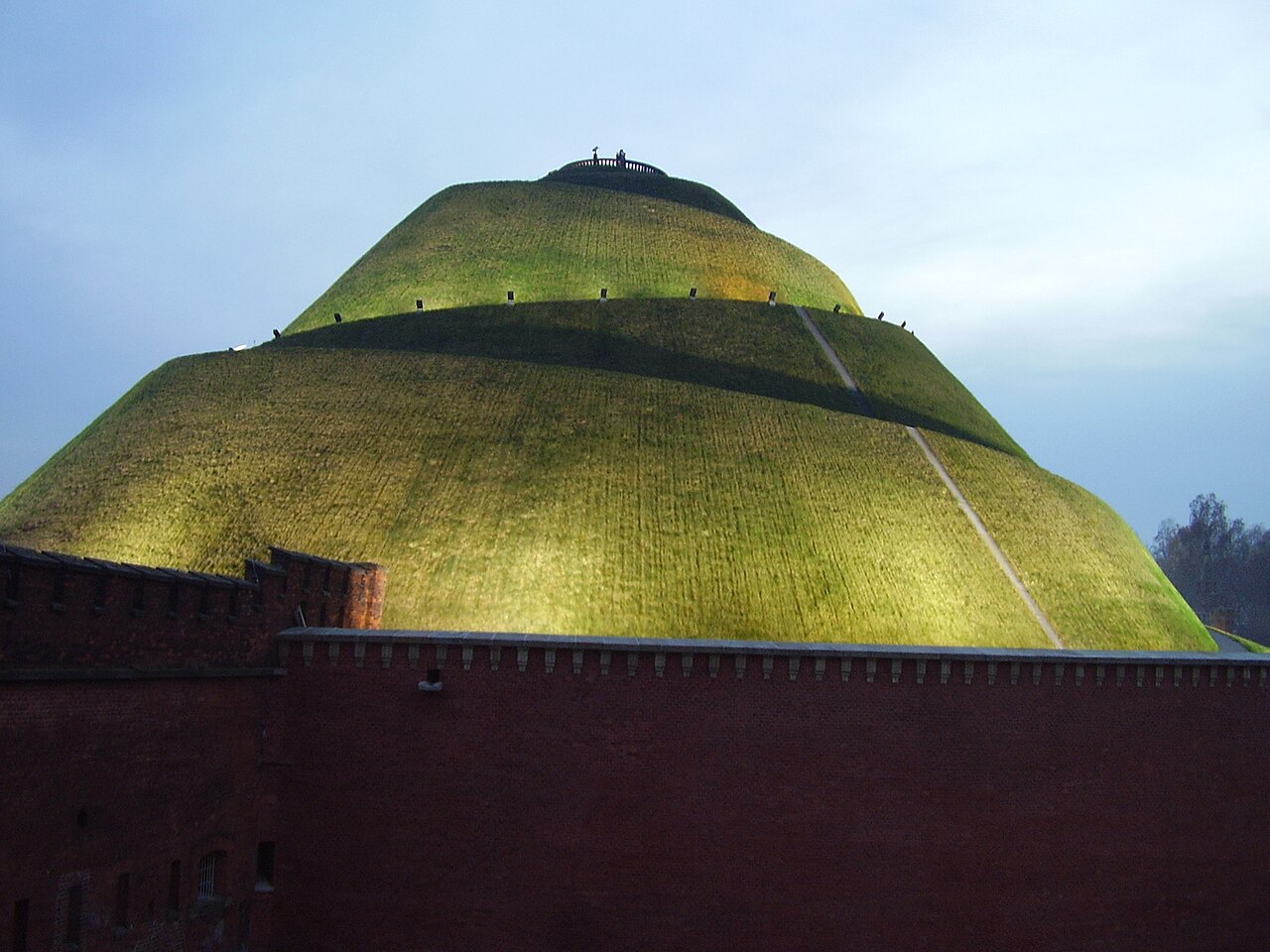 Kościuszko Mound in Kraków at dusk: a large grassy artificial hill lit from below, rising behind the red-brick wall of the fortress that surrounds its base