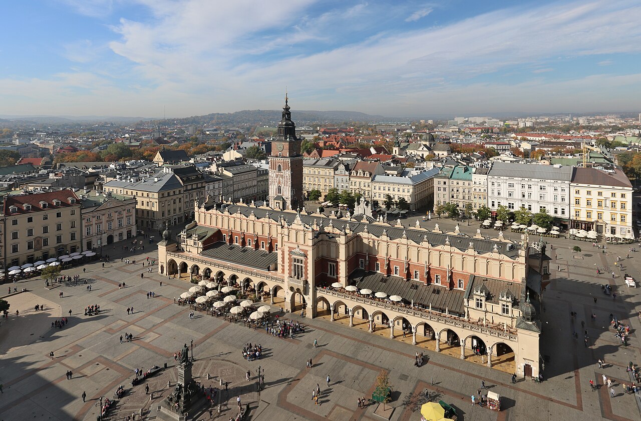 Kraków's Main Market Square from above: the long arcaded Cloth Hall in the middle, the town hall tower beside it, café umbrellas around the edges and the city rooftops beyond