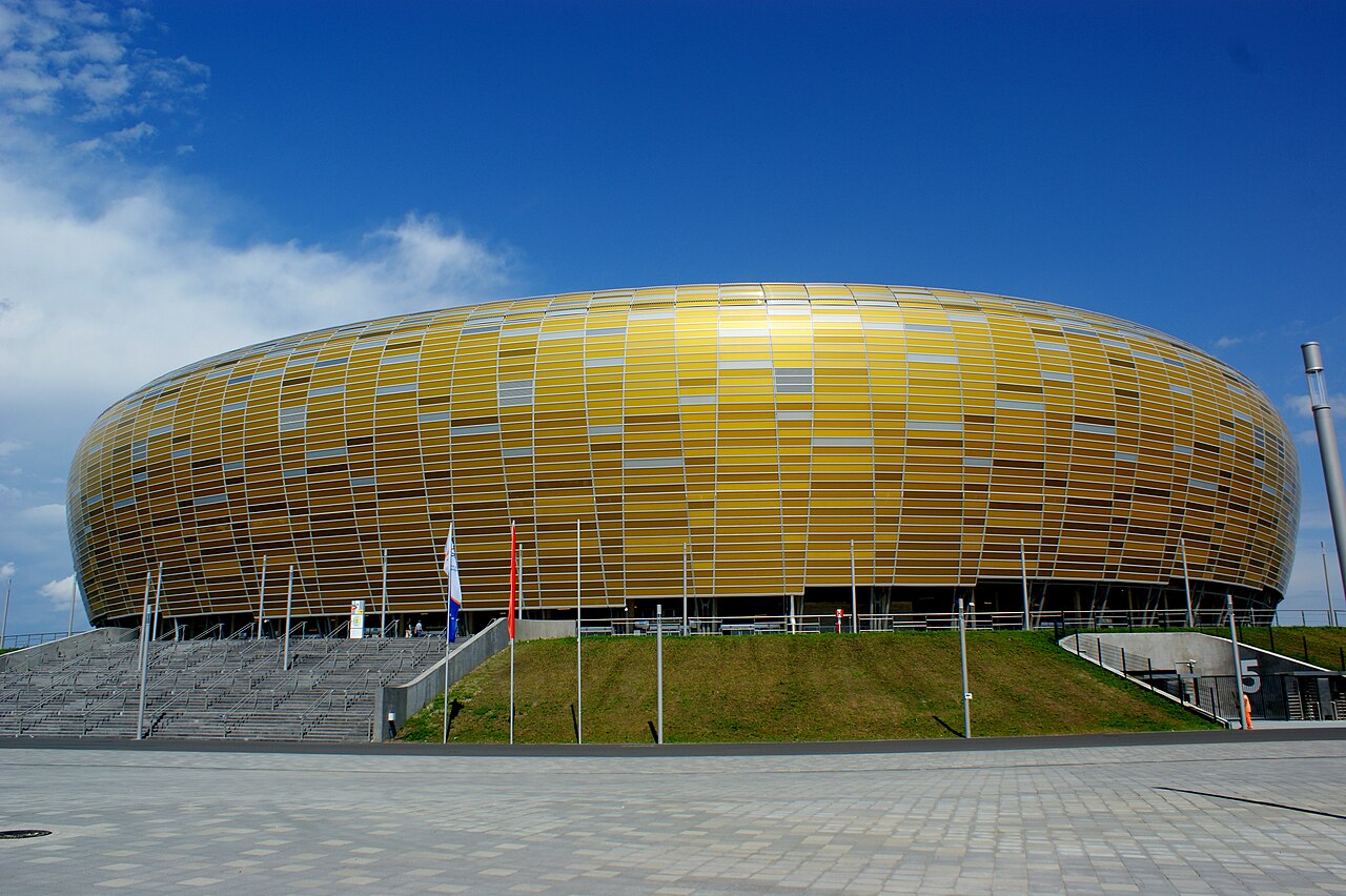 The stadium in Gdańsk seen from outside on a clear day: a low oval clad in overlapping amber and gold panels rising from a grass bank, with flagpoles and a wide paved forecourt in front
