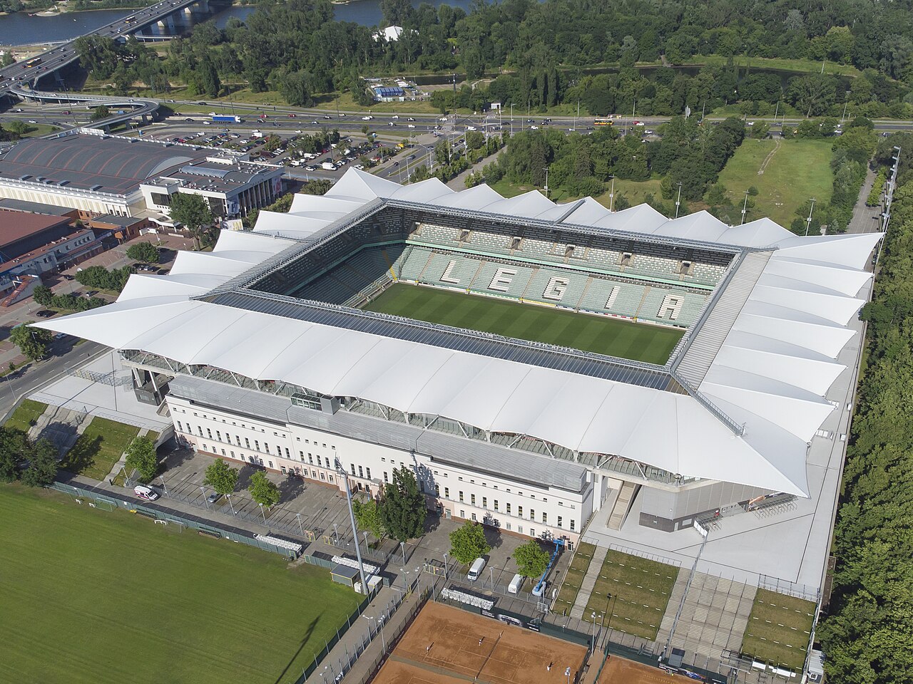 Stadion Wojska Polskiego seen from the air: four covered stands around a green pitch, LEGIA spelled out in the seats of the far stand, with the Vistula and Warsaw traffic behind