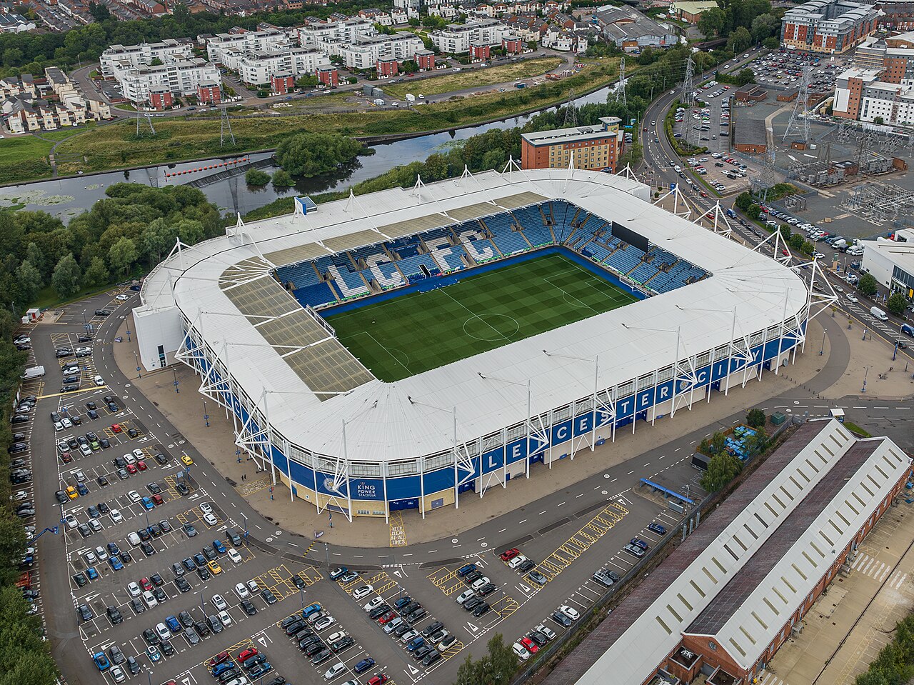 The King Power Stadium from the air: a modern enclosed blue and white bowl with LCFC picked out in the seats and LEICESTER CITY along the outside of the roof, standing beside the River Soar with large car parks around it