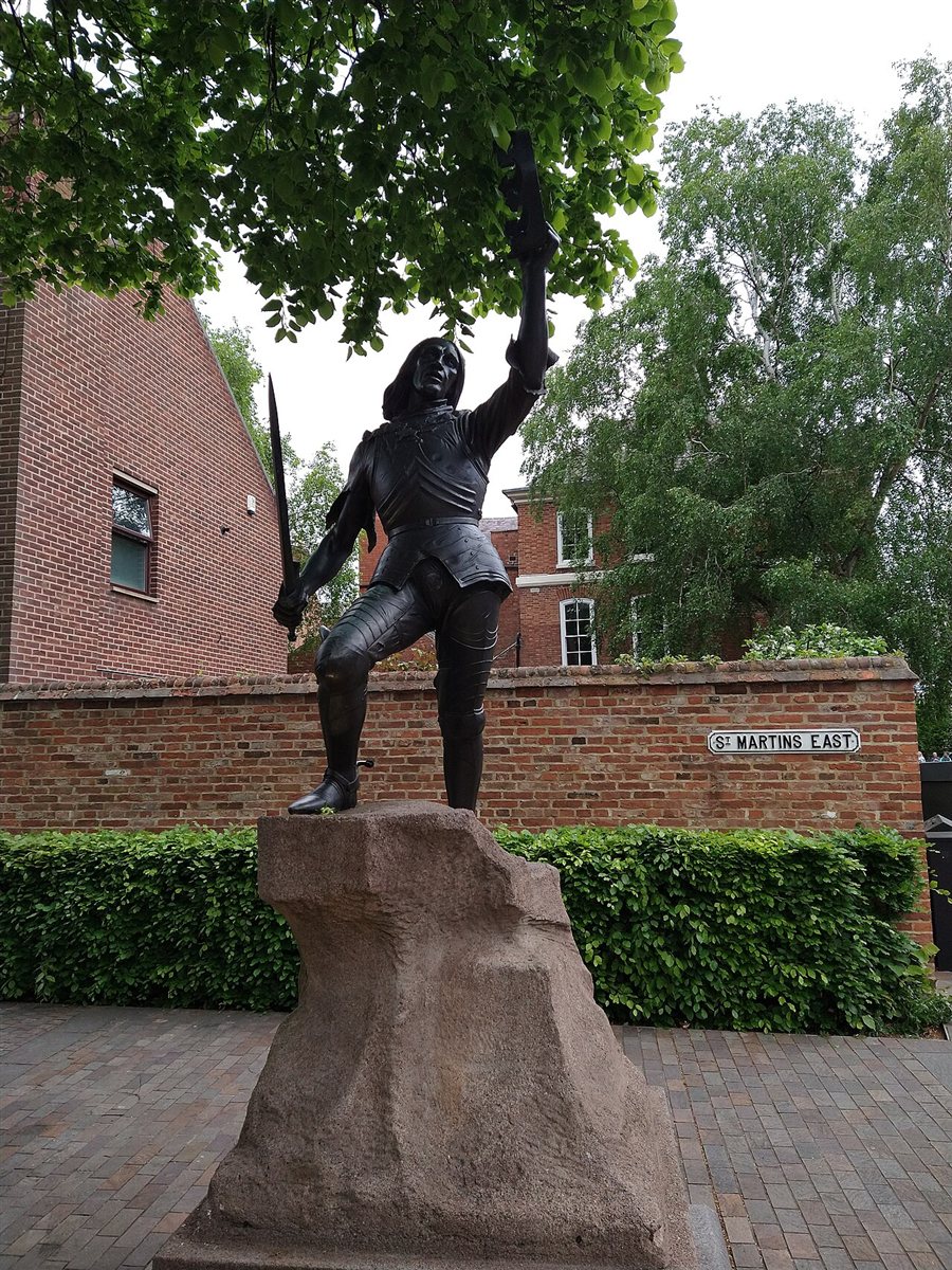 The bronze statue of Richard III in Leicester: a figure in plate armour standing on a rough stone plinth, holding a sword low in one hand and raising a crown above his head with the other, with a brick wall and trees behind