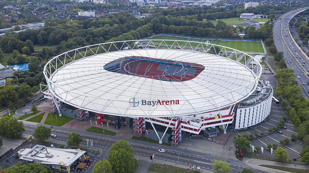 The BayArena from the air: a fully enclosed oval stadium under a white membrane roof lettered BayArena, with a curved white hotel block attached to one side, training pitches behind it and a motorway running past