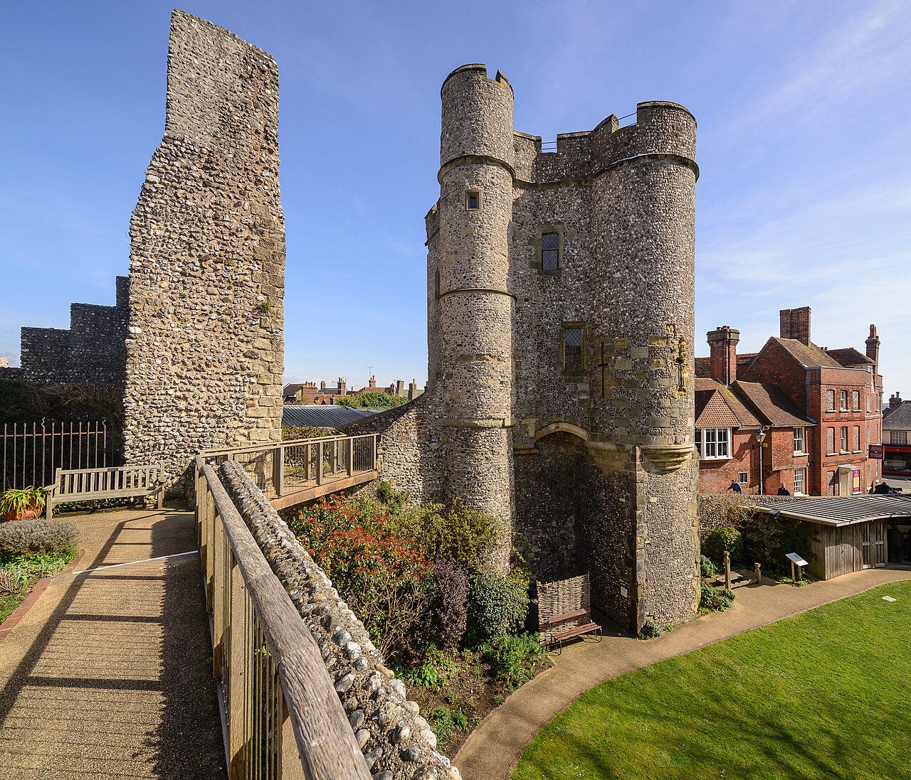 Lewes Castle: a Norman gatehouse of grey flint with two tall round towers flanking an arched entrance, a ruined section of curtain wall to the left, a wooden walkway and shrubs in front, and red-brick town buildings immediately beyond