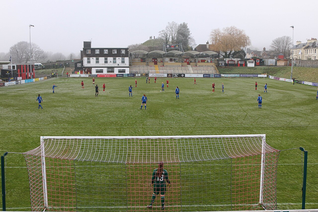 A women's match in progress at the Dripping Pan seen from behind the goal: players in red and blue on a frosty pitch, a small covered stand and white clubhouse along the far side, a grassy bank rising behind them with a few spectators on the mound, and mist over the hills