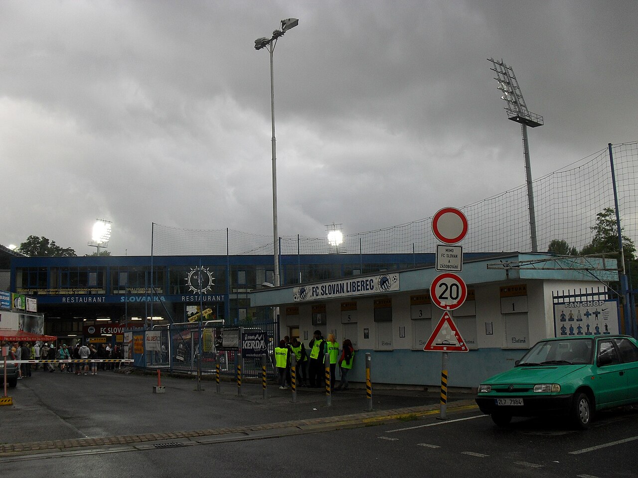 The outside of Stadion u Nisy on a wet evening: a low blue and white building with numbered ticket windows and an FC SLOVAN LIBEREC sign, stewards in high-visibility jackets, floodlights lit above