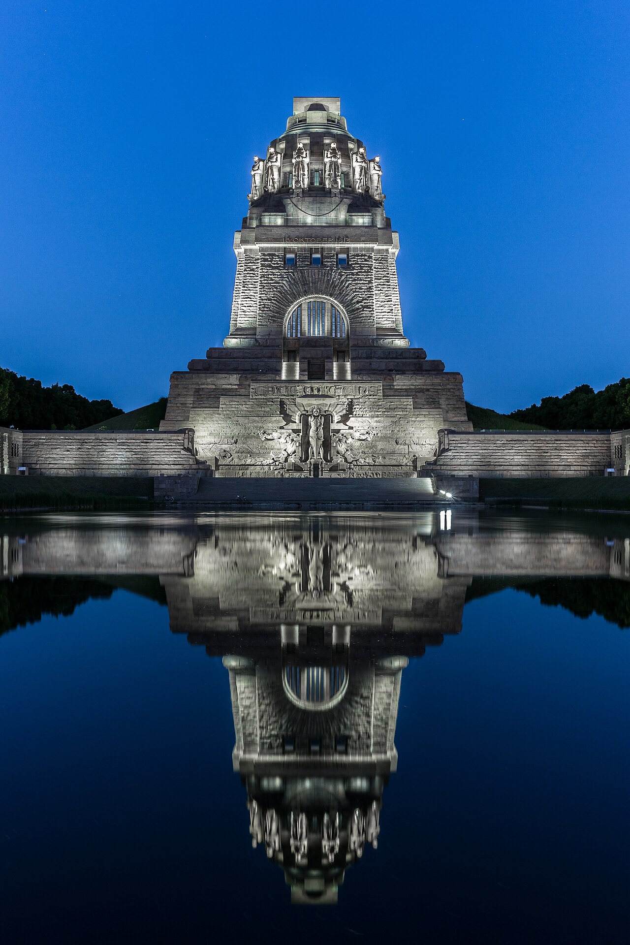 The Monument to the Battle of the Nations in Leipzig, floodlit at night: a colossal stepped stone tower with giant guardian figures near the top, reflected in the long still pool in front of it