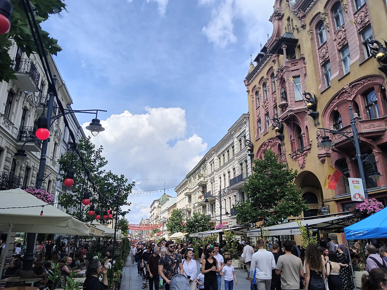 ul. Piotrkowska in Łódź full of people on a summer day: ornate nineteenth-century tenement facades on both sides, café terraces under awnings and red lanterns strung above the street