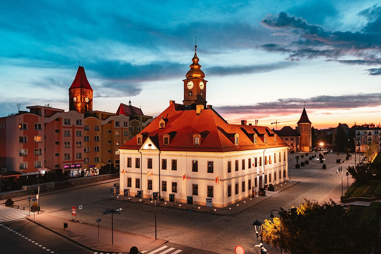 Lubin's town hall floodlit at dusk: a white building with a red roof and a clock tower on a paved square, an old church tower and a stone defensive tower behind, post-war blocks of flats to the left