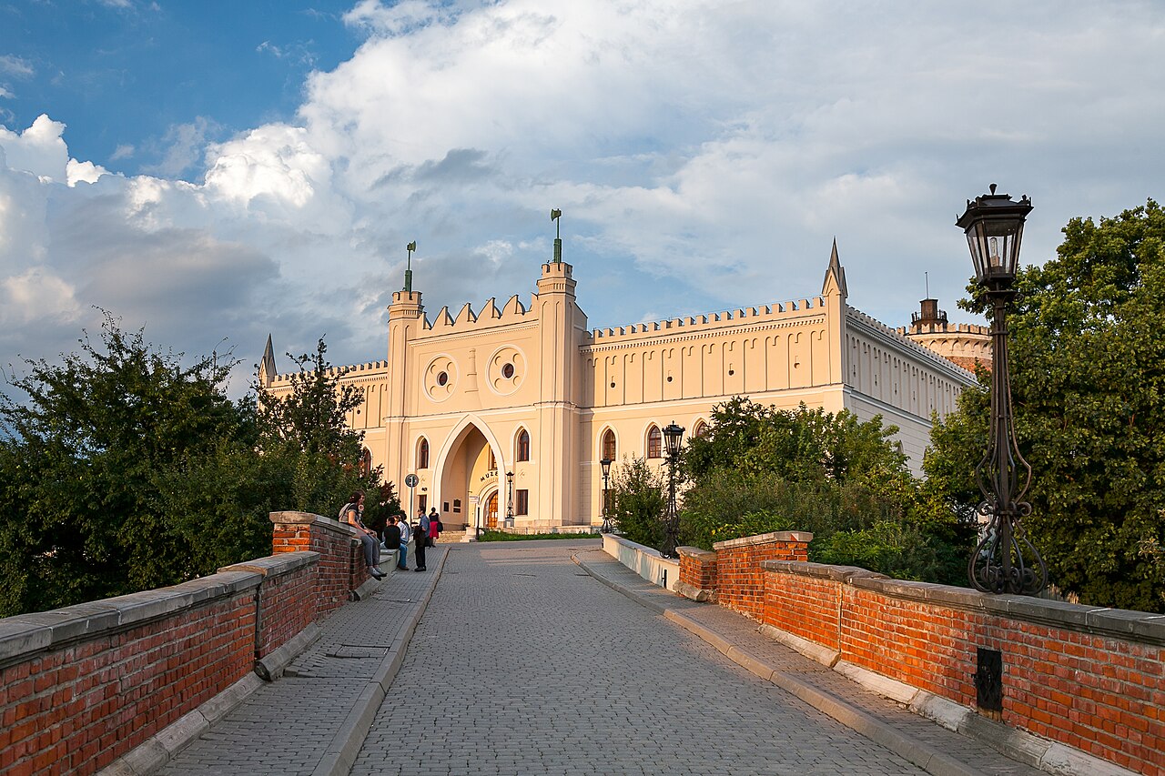 Lublin Castle at the end of a cobbled ramp with red-brick parapets and an ornate lamp post: a cream neo-Gothic gatehouse with crenellations and a pointed arch, under an evening sky