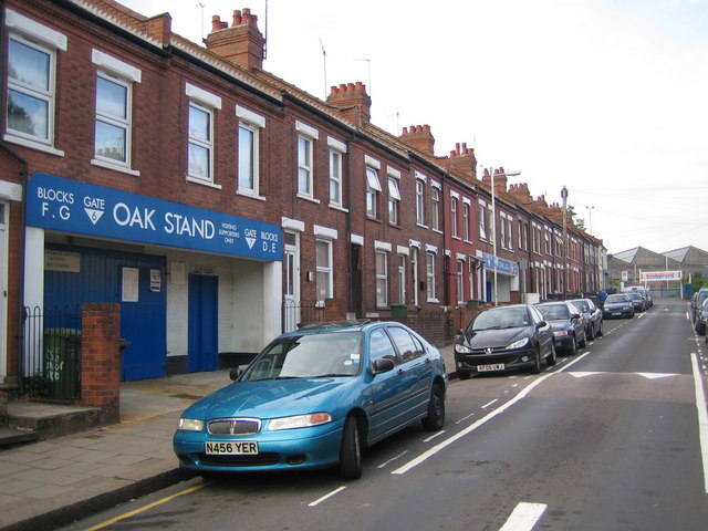 The away entrance at Kenilworth Road: a row of Victorian red-brick terraced houses on an ordinary residential street, with a blue and white football turnstile entrance built into the middle of the terrace reading OAK STAND, BLOCKS F G, VISITING SUPPORTERS ONLY, and cars parked at the kerb outside