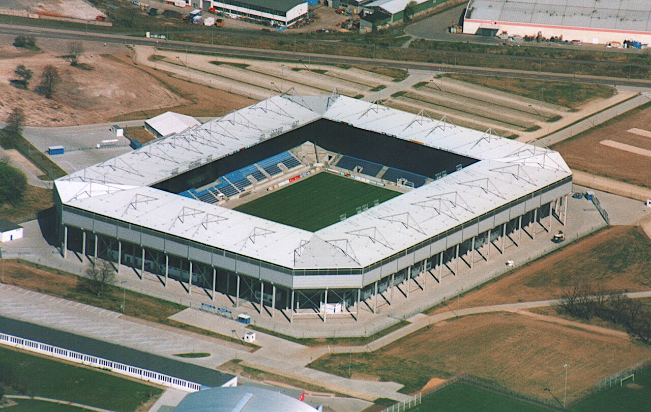 The Magdeburg stadium from the air: a square football-specific ground with white roofs over all four stands, blue seats inside, no running track, standing on open ground beside a road
