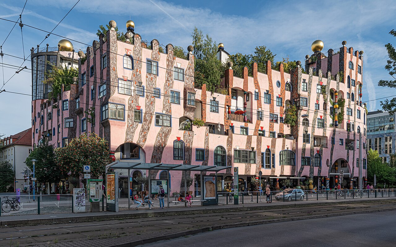 The Green Citadel in Magdeburg: a long pink apartment building with wavy stone columns, irregular windows of different sizes, golden spheres on the roofline and trees growing out of the upper storeys, with tram wires crossing in front