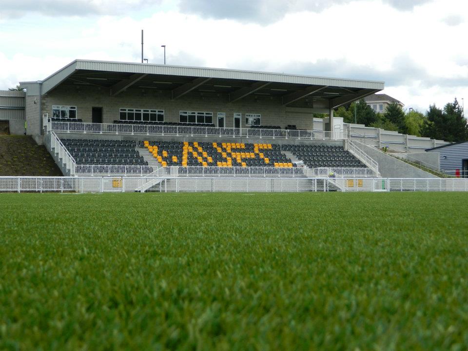 The main stand at the Gallagher Stadium seen across the pitch: a single covered stand of black and amber seats above a pale stone clubhouse with white railings, and in the foreground the uniform bright green of an artificial playing surface