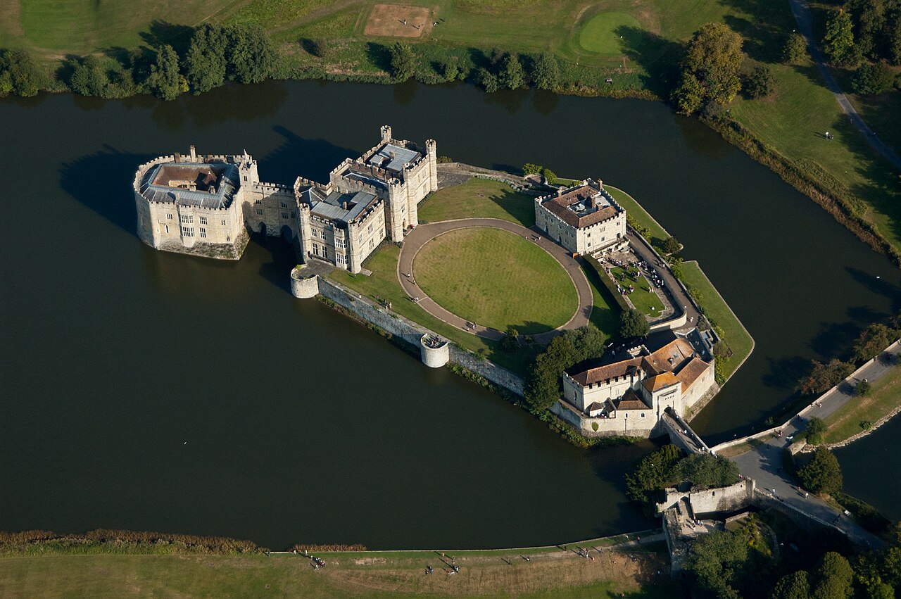 Leeds Castle from the air: a pale stone castle with battlemented towers standing on two islands in a broad lake, joined by a bridge, with a circular lawn inside the walls and parkland and woodland all around
