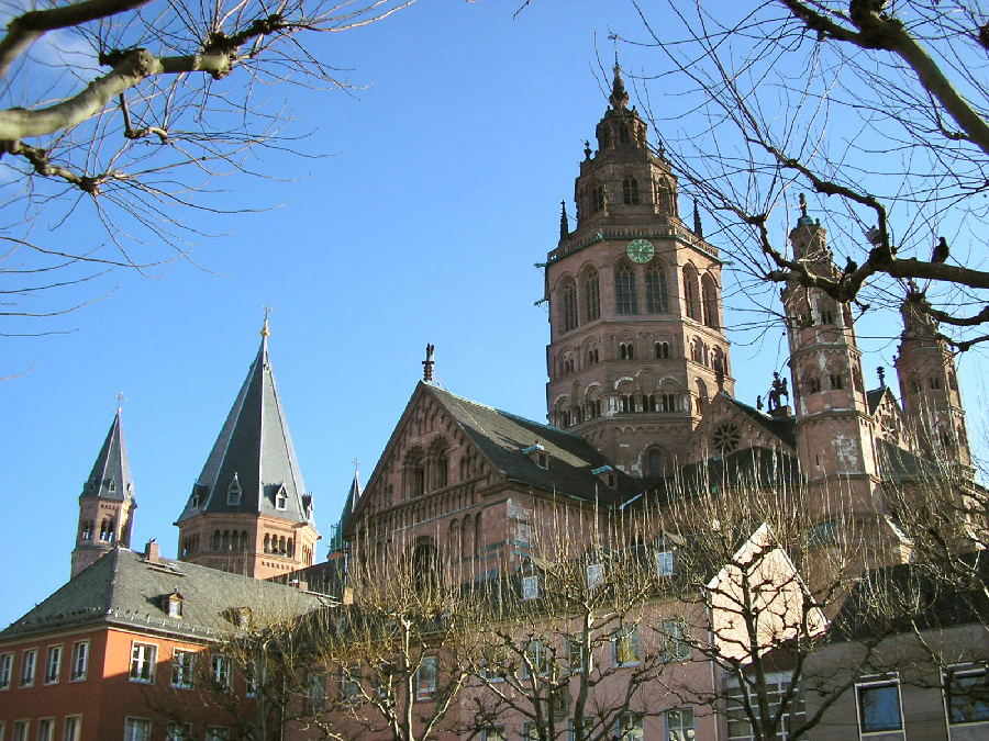 Mainz cathedral rising above the rooftops: a red sandstone Romanesque church with a tall octagonal crossing tower, a clock face, several spires and bare winter trees in front of it