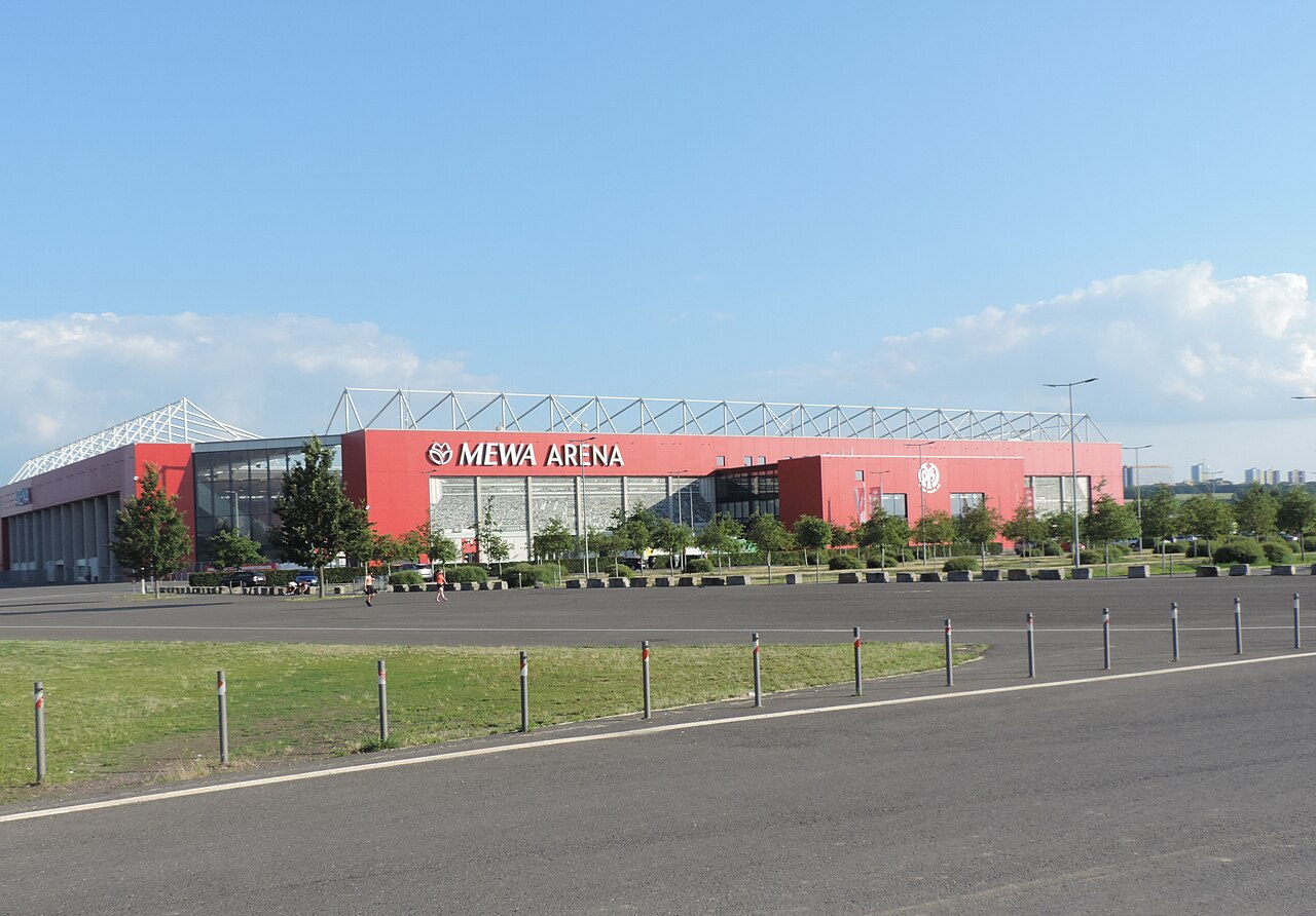 The MEWA Arena seen from across a vast empty car park: a long red-clad stand with MEWA ARENA lettering and an exposed white steel roof truss, open fields and tower blocks on the horizon