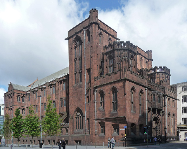 The John Rylands Library on Deansgate in Manchester: a large red sandstone neo-Gothic building with pinnacled towers, pointed arched windows and an elaborately carved entrance, standing on a city street with young trees in front