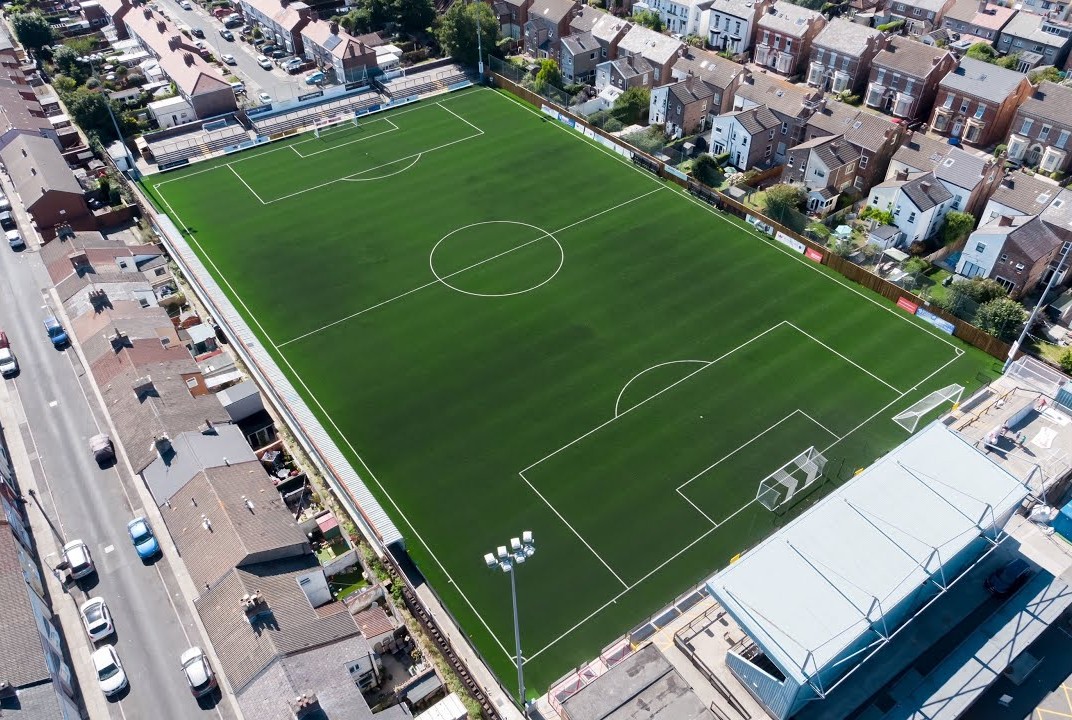 Rossett Park from a drone: a green pitch filling the frame with only narrow strips of stand around it, and the back gardens and roofs of terraced and semi-detached houses pressed directly against three sides of the ground, with a residential street and parked cars along the fourth