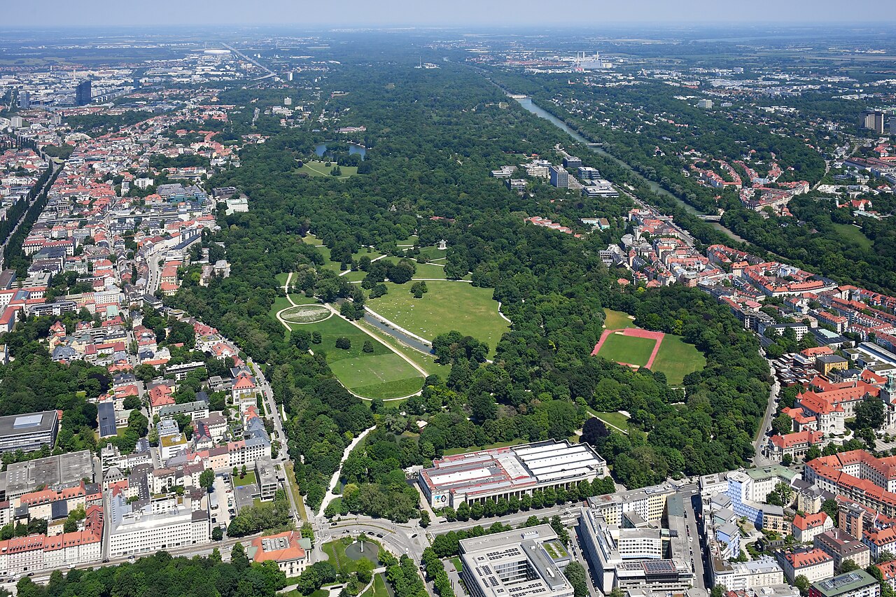Aerial view of the Englischer Garten in Munich: a vast wedge of parkland with meadows, woods and a lake running from the edge of the city centre out towards the horizon, dense streets on both sides