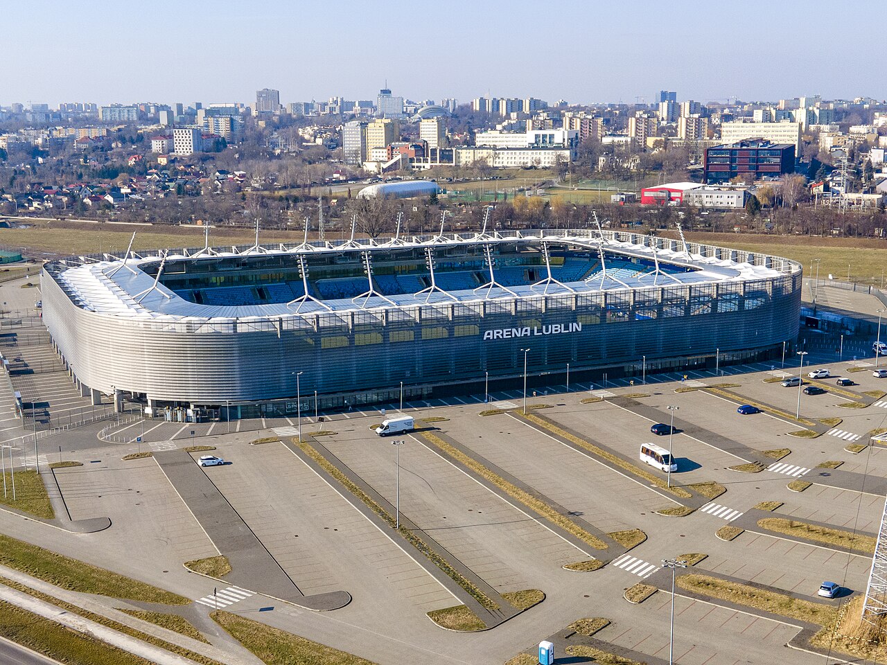 Arena Lublin from the air: an oval stadium clad in silver vertical louvres with ARENA LUBLIN written across the side, blue seats visible inside, a large empty car park in front and the Lublin skyline behind