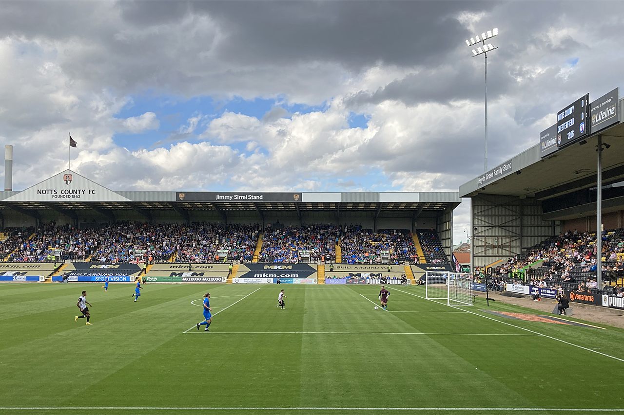 A match in progress at Meadow Lane: players in black and white stripes against opponents in blue, a covered stand opposite with the lettering NOTTS COUNTY FC FOUNDED 1862 and Jimmy Sirrel Stand along its roof, and a crowd beneath it under a cloudy sky