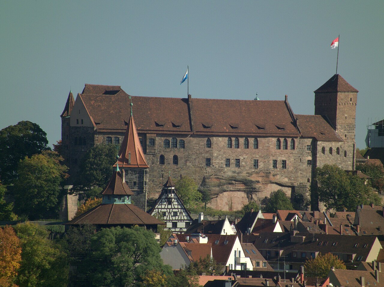 The Kaiserburg in Nuremberg seen from across the old town: a long stone and half-timbered castle standing on a bare sandstone outcrop, with a round tower and a square keep flying flags, and steep red-tiled roofs of the old town below it