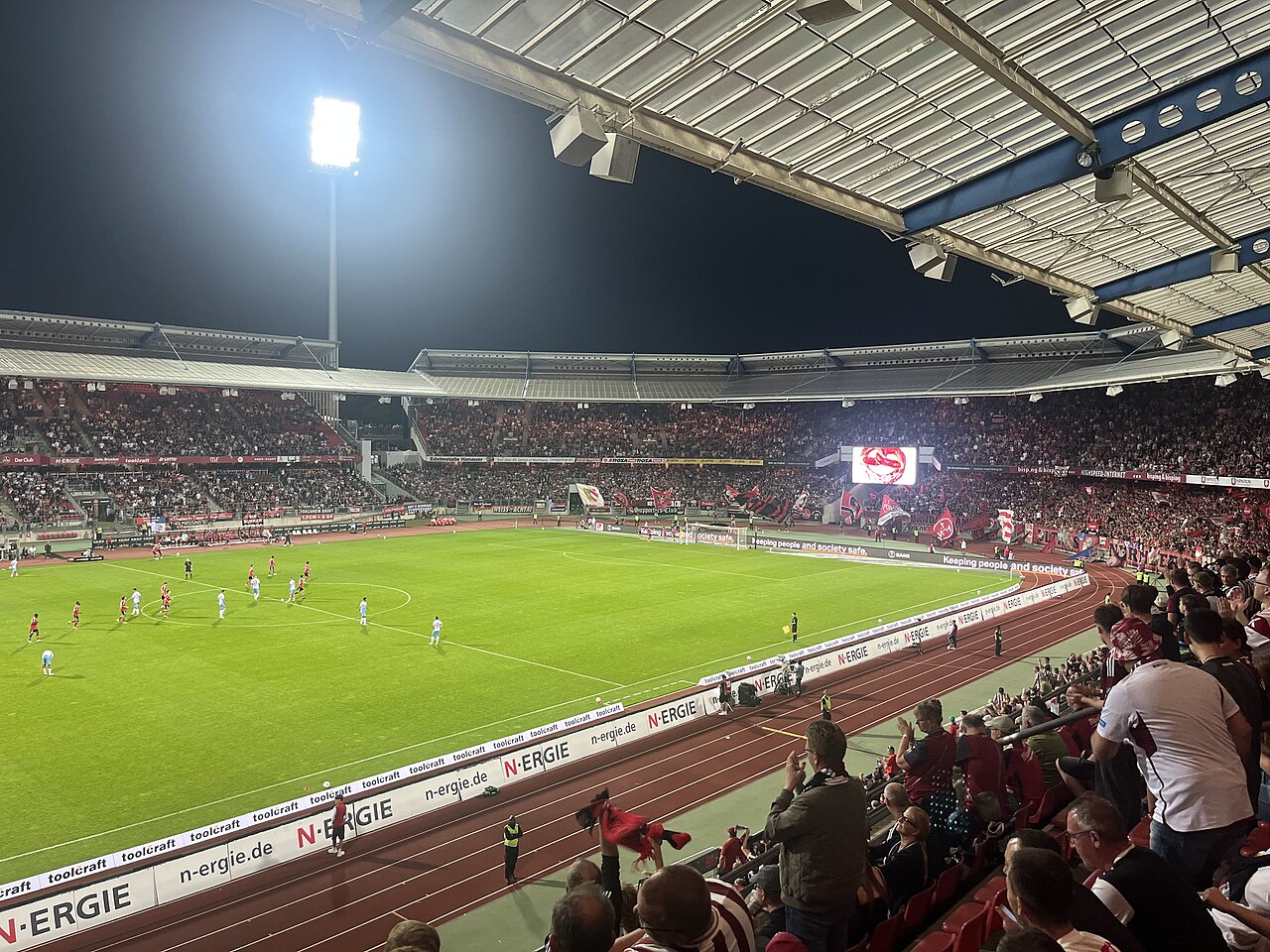 The Max-Morlock-Stadion during a floodlit evening match, seen from a side stand: a red running track separating the crowd from the pitch, packed tiers of supporters around the bowl and flags waving in the home end