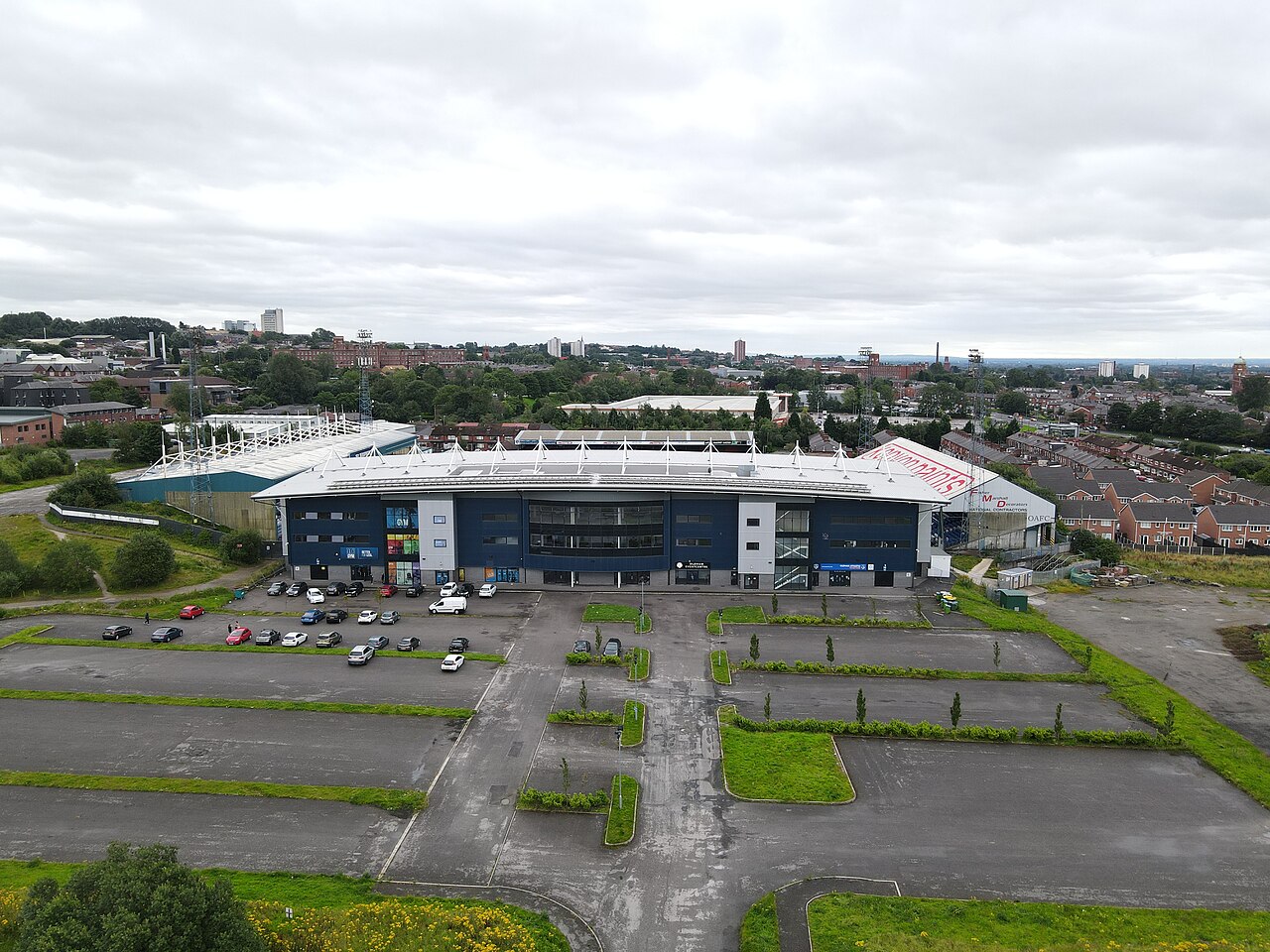 Boundary Park from the air under a heavy grey sky: a blue and white stadium with a white roof, surrounded by very large mostly empty car parks and scrubby open ground, with the mill chimneys and rooftops of Oldham spreading away behind it