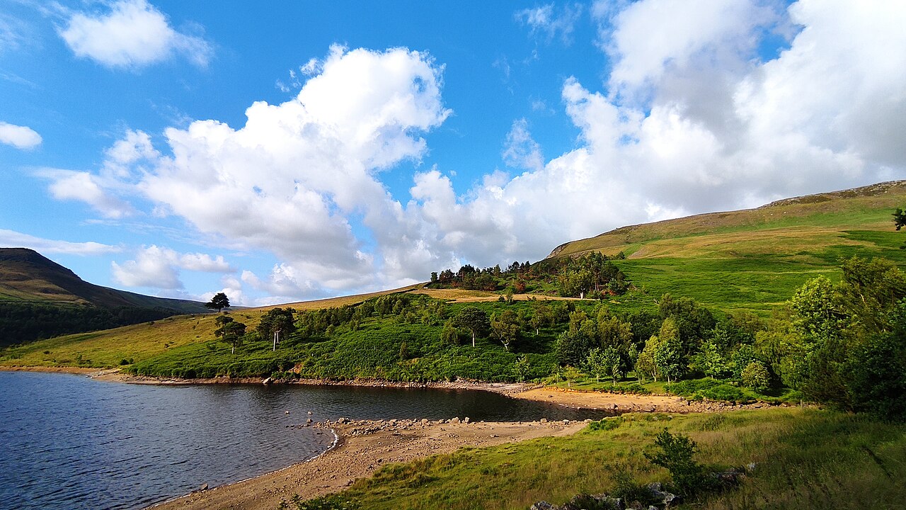 Dovestone Reservoir under a blue sky with white clouds: dark water in the foreground, a shingle shore, wooded slopes rising to bare green moorland, and the flat edge of a Pennine hill on the horizon