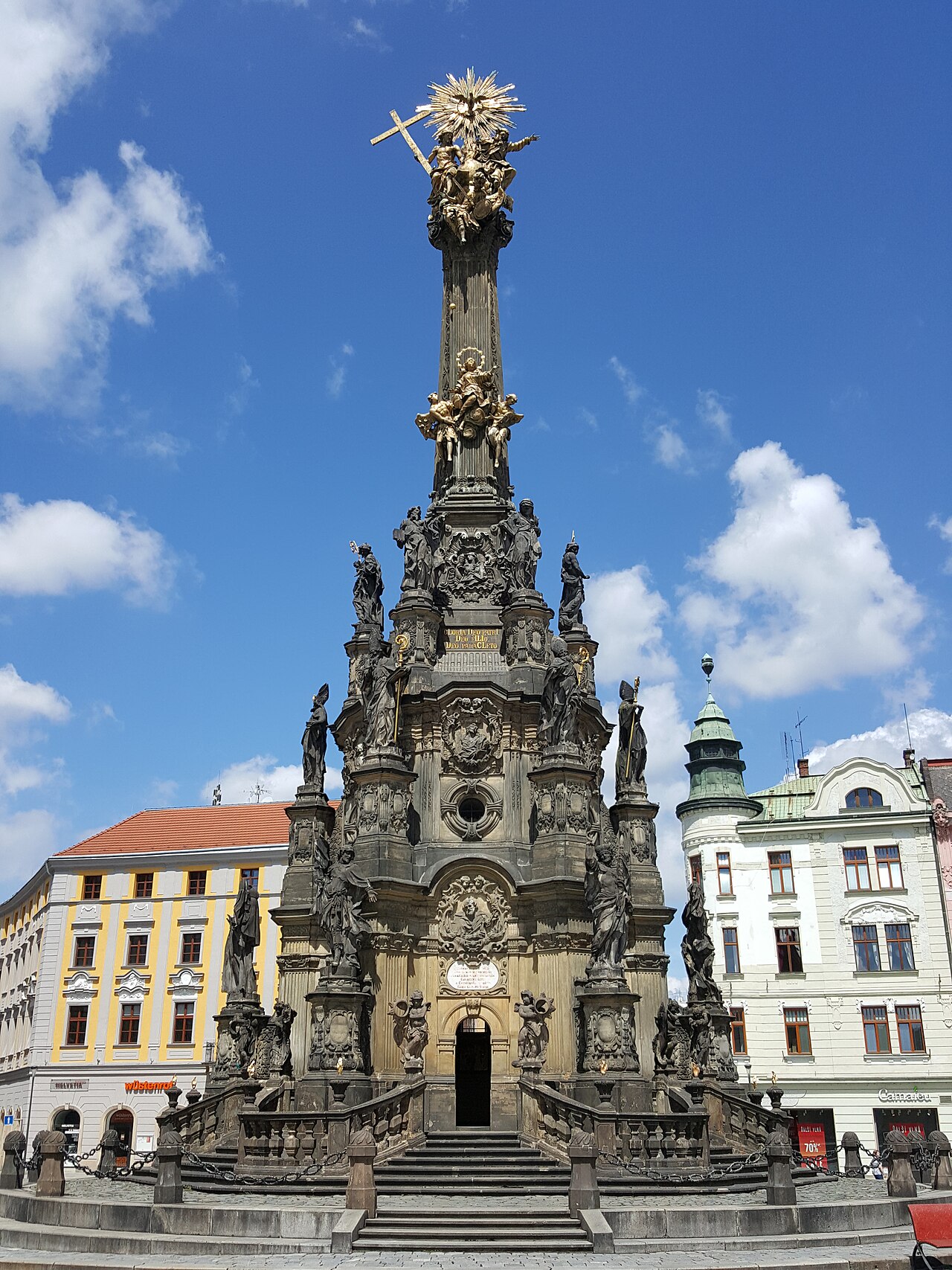 The Holy Trinity Column in Olomouc: a towering baroque monument of dark stone crowded with saints, topped by gilded figures and a golden sunburst, standing on the town square against a blue sky