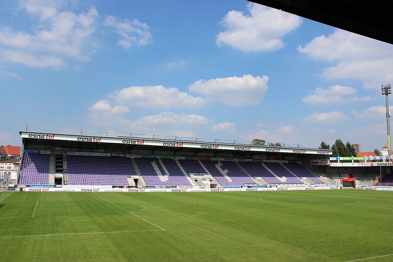 Inside the Stadion an der Bremer Brücke: a single covered stand of purple seats with a sponsor name along its roof edge, advertising hoardings around the pitch, a floodlight pylon at the corner and the roofs of ordinary houses visible just beyond the stand