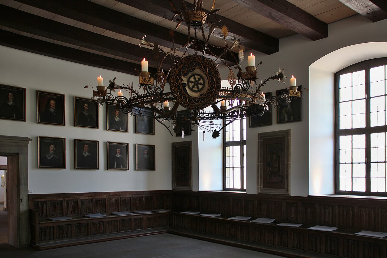 The Hall of Peace in Osnabrück town hall: a white-walled room with a dark beamed ceiling, two rows of small framed portraits of seventeenth-century delegates along the walls, carved wooden benches around the edges and a large wrought-iron candle chandelier hanging in the centre
