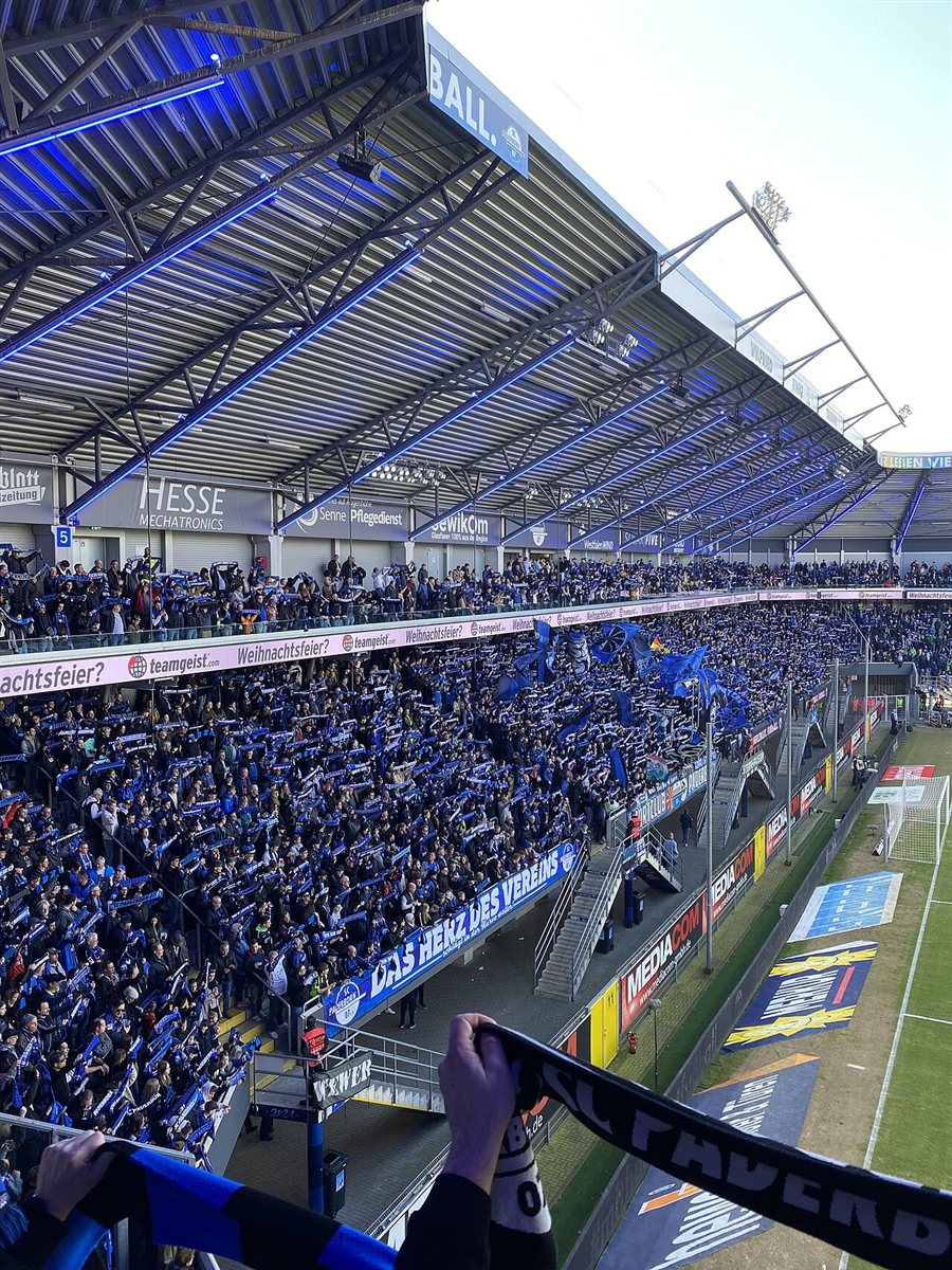 Inside the Home Deluxe Arena: a packed two-tier stand of Paderborn supporters holding blue and white scarves above their heads, blue strip lighting along the roof girders and a banner reading Das Herz des Vereins along the front of the terrace