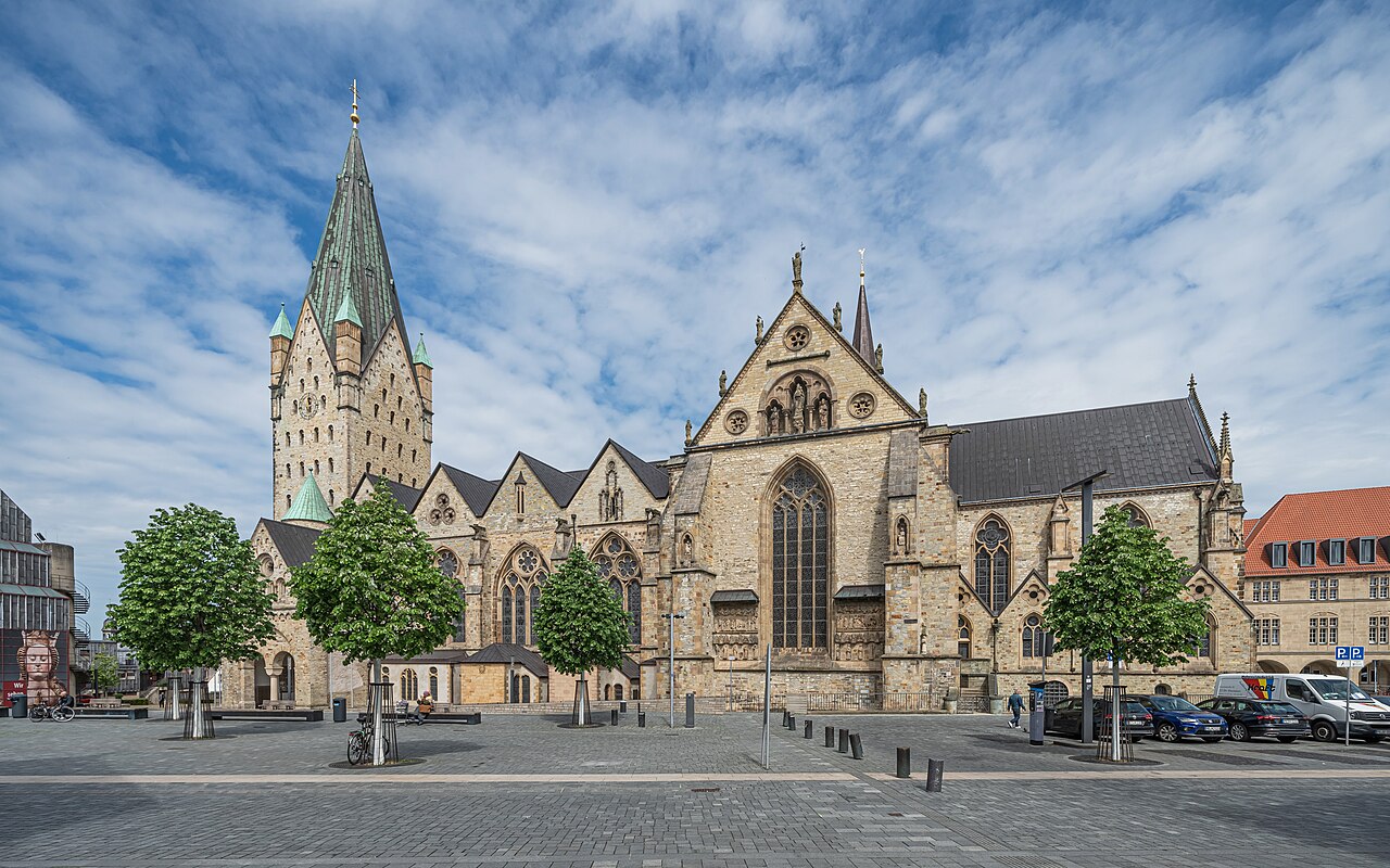 Paderborn cathedral seen across an open paved square: a long pale sandstone church with a single square tower topped by a green copper spire, tall gothic windows and young trees along the front