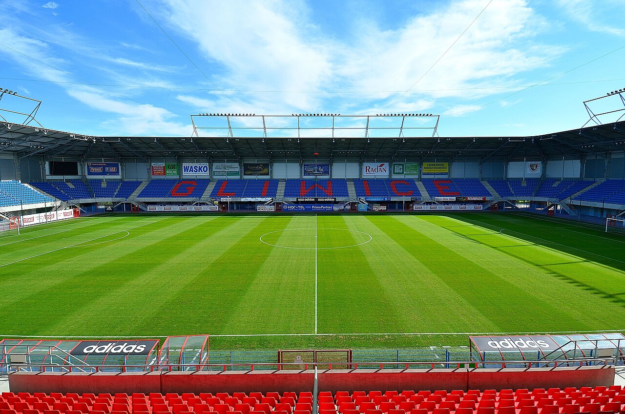 The inside of Piast Gliwice's stadium seen from behind a goal: a striped green pitch, a covered main stand opposite with GLIWICE spelled out in red letters across the blue seats